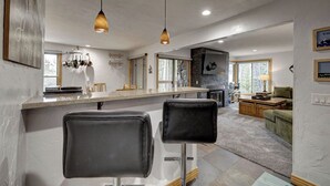 A modern kitchen with a central island, bar stools, pendant lighting, and a stone fireplace. - Large Corner 2 Bedroom Unit Mountain Vibes (Breckenridge)