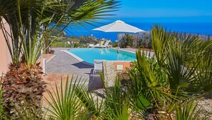 A pool area with a view of the sea, surrounded by palm trees and a white umbrella. - VILLA TARI' (Scopello)