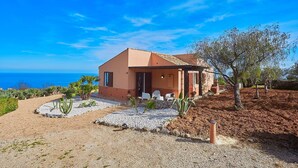 A single-story house with a sloped roof, a stone pathway leading to the entrance, and a view of the sea. - VILLA TARI' (Scopello)