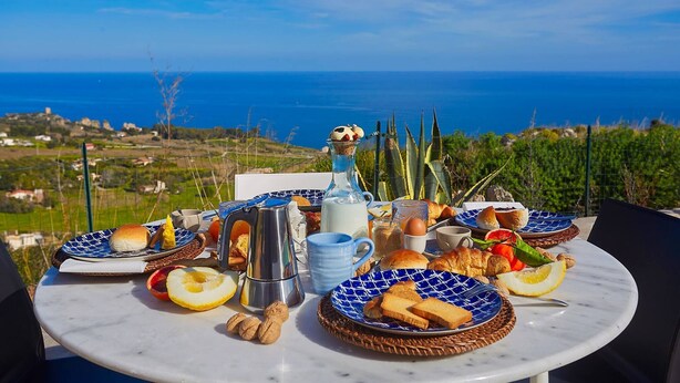 A table set with breakfast items including bread, eggs, and fruits, overlooking a scenic coastal view. - VILLA TARI' (Scopello)