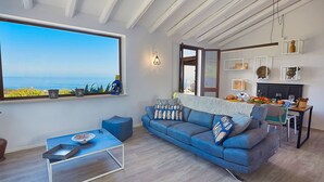 A modern living room with a large window, a blue sofa, a small table, and a dining area with a fireplace. - VILLA TARI' (Scopello)