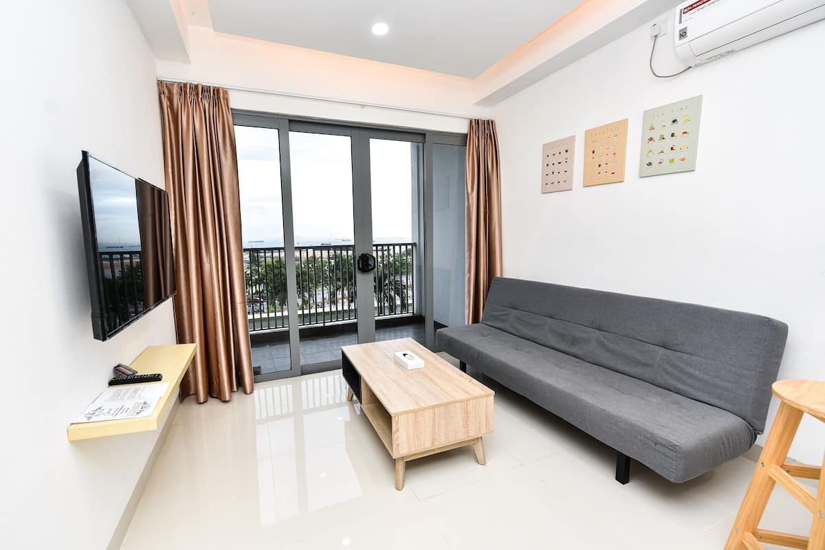 Ein modernes Wohnzimmer mit grauem Sofa, einem Holztisch, einem Flachbildfernseher und einem Balkon mit Vorhängen.