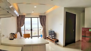 A modern living room with a large window, a flat-screen TV, a coffee maker, and a refrigerator. - Lovina Residences at Harbour Bay (Batam)