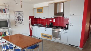 Cocina pequeña con armarios blancos, salpicadero rojo y una estufa. Se ve una mesa de comedor con sillas y un televisor en un estante.