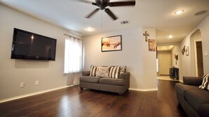 A living room with a flat-screen TV, a gray sofa, wooden flooring, and a ceiling fan.