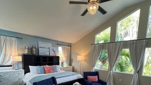 A spacious bedroom with a large bed, two armchairs, a round ottoman, and a ceiling fan.