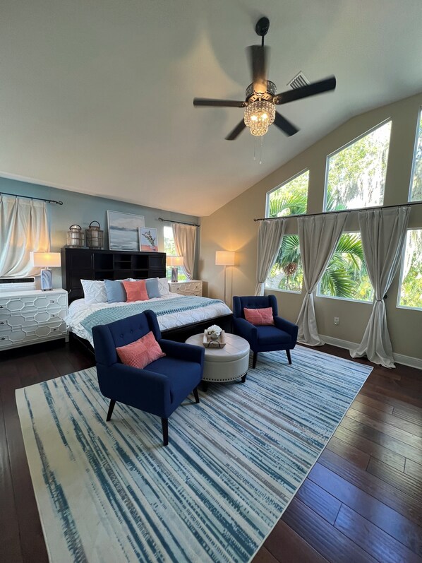 A spacious bedroom with a large bed, two armchairs, a round ottoman, and a ceiling fan.