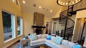 A spacious living room with a sectional sofa, a coffee table, and a fireplace. There is a staircase with a wrought iron railing leading to an upper level.
