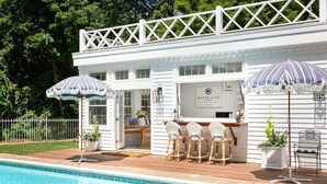 Eine Poolbar mit Sitzgelegenheiten und Sonnenschirmen, ein weißes Gebäude mit dem Schriftzug "Manor Lane" und ein umzäunter Hinterhof.