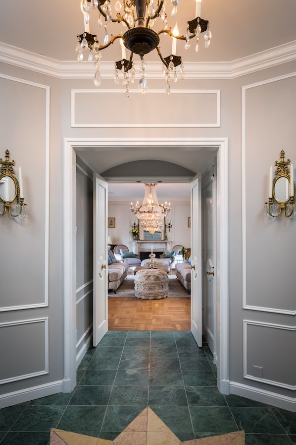 A hallway with a chandelier, a mirror, and a view into a living room with a fireplace.