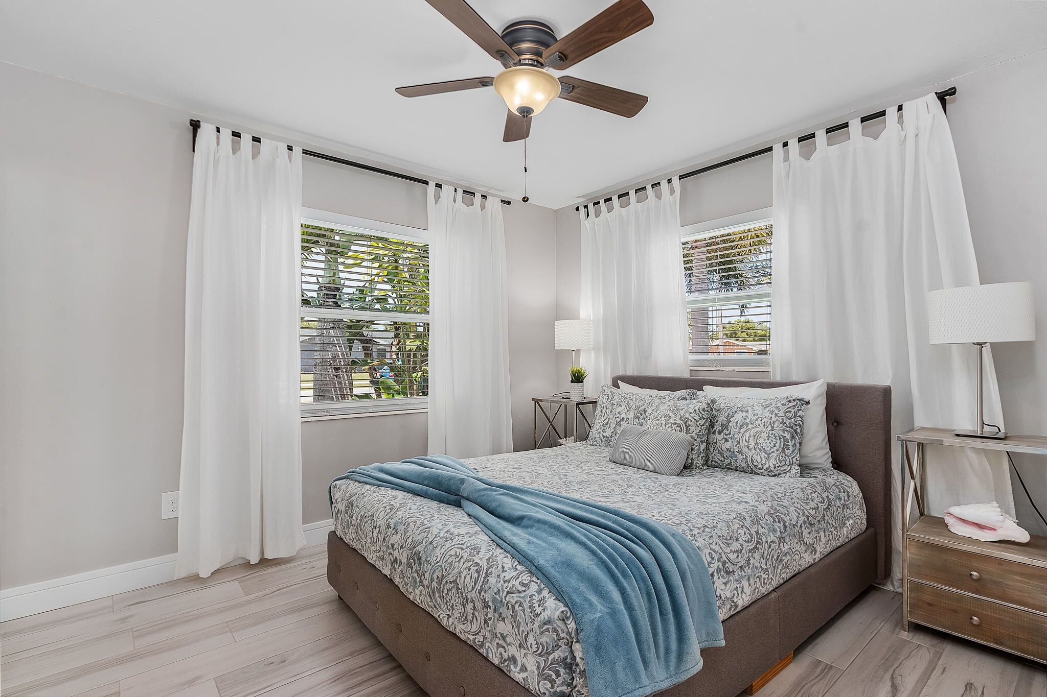 A bedroom with a bed, curtains, a ceiling fan, a bedside table with a lamp, and a window with blinds.