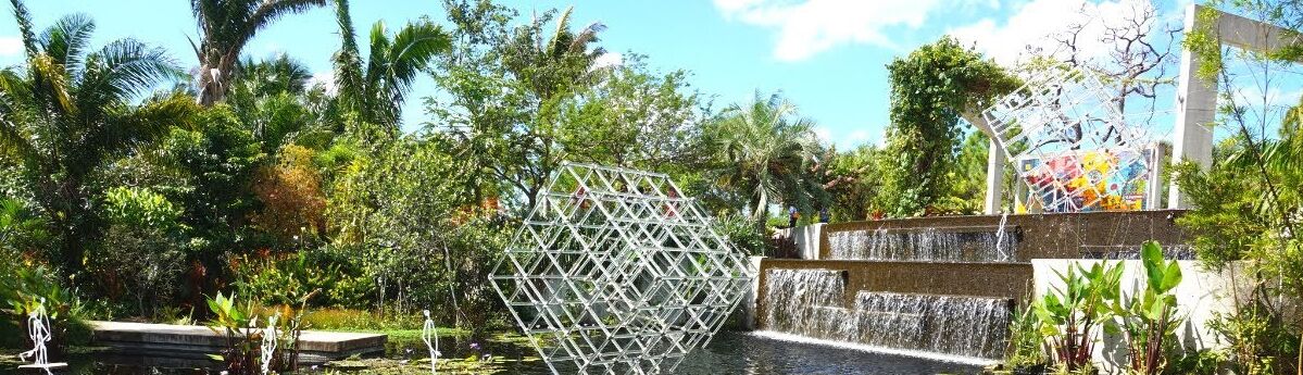A geometric metal sculpture in a lush garden with a waterfall and a stone structure.