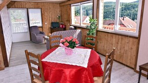 A cozy room with wooden walls, a dining table with a red tablecloth, and a chair. There is a window with a view of houses and trees.