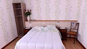 A bedroom with a wooden bed, a nightstand, a chair, and a shelf with a vase of flowers.