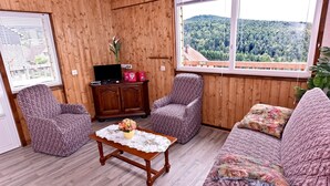 A cozy room with wooden walls, a large window with a forest view, two patterned armchairs, a wooden coffee table with a lace tablecloth, and a cabinet with electronic devices.