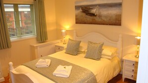 A neatly made bed with pillows and folded towels, a painting of a boat on the wall, and a window with curtains. - Elm Cottage, Purpose Built Wheelchair Friendly Cottage (Truro)