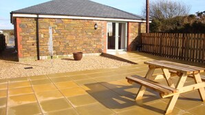 A stone building with a tiled patio, a wooden bench, and a brown pot. - Elm Cottage, Purpose Built Wheelchair Friendly Cottage (Truro)