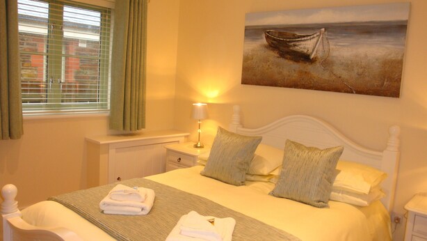 A neatly made bed with pillows, a bedside lamp, and a painting of a boat on the wall. - Elm Cottage, Purpose Built Wheelchair Friendly Cottage (Truro)