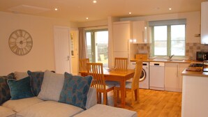 A compact living space with a dining area, a sofa with patterned cushions, a wooden dining table, and a kitchen with a dishwasher and microwave. - Elm Cottage, Purpose Built Wheelchair Friendly Cottage (Truro)