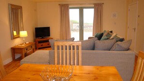 A living room with a wooden table, a sofa, a television, and a view of the outdoors. - Elm Cottage, Purpose Built Wheelchair Friendly Cottage (Truro)