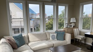 A living room with a sectional sofa, a coffee table, a desk, and a chair. Large windows provide views of the neighborhood.