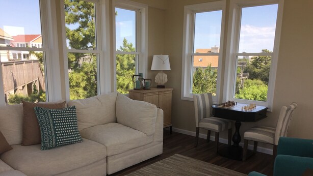 A living room with a beige sofa, teal armchairs, a small table with chess pieces, and large windows offering a view of trees and buildings.