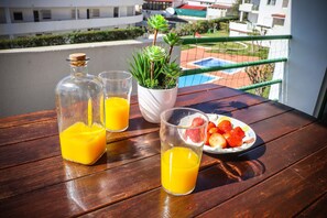 Una mesa de madera con una botella de jugo de naranja, un vaso de jugo de naranja y un plato de fresas y otras frutas en un balcón con vista a una piscina y edificios.