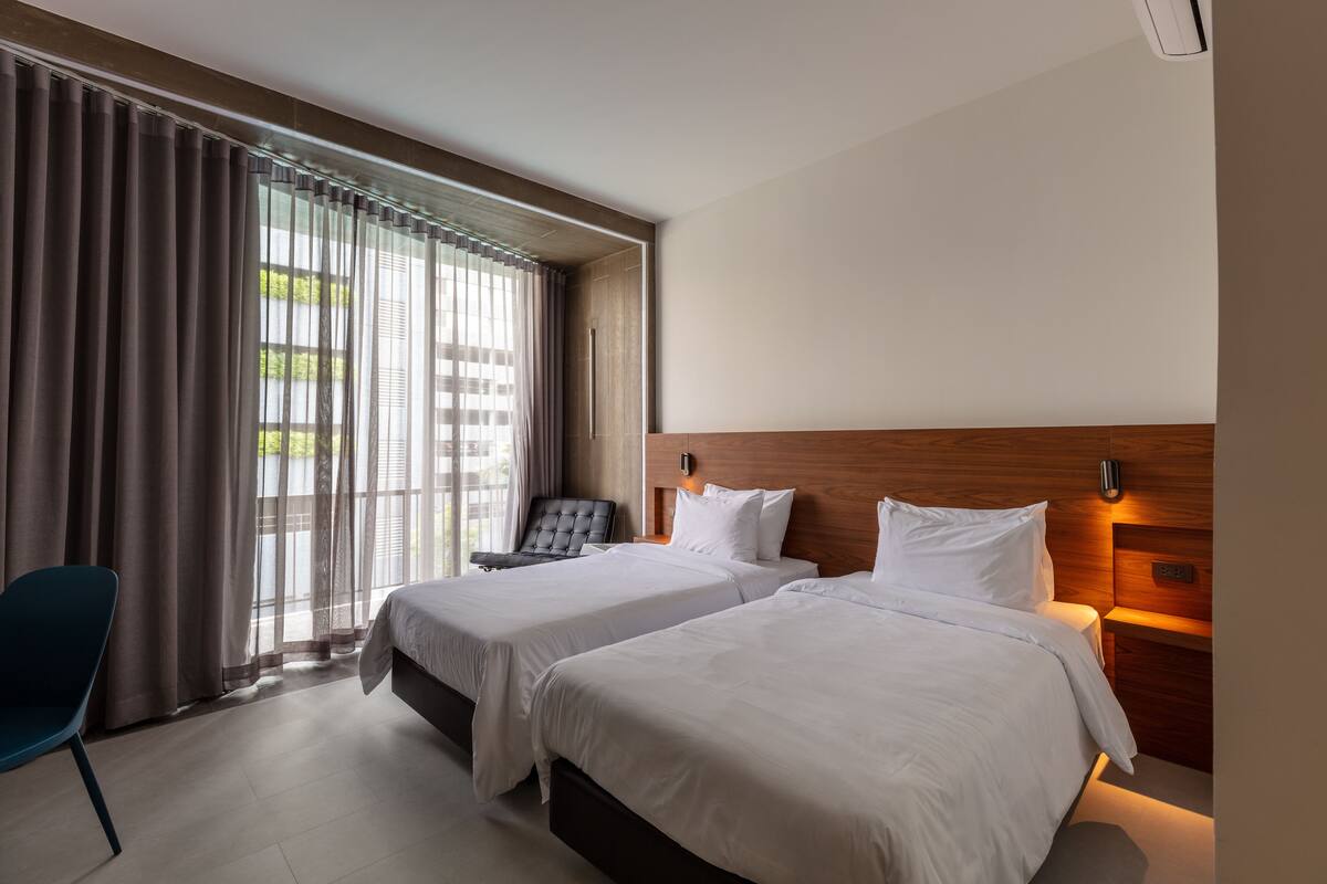 A hotel room with two beds, a blue chair, and a view of greenery through the window.