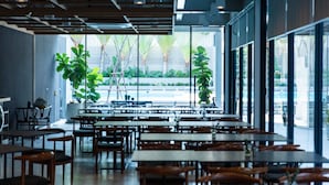 A spacious indoor dining area with multiple tables and chairs, large windows offering views of the outside, and potted plants adding greenery. - Reno Hotel Bangkok (Bangkok)