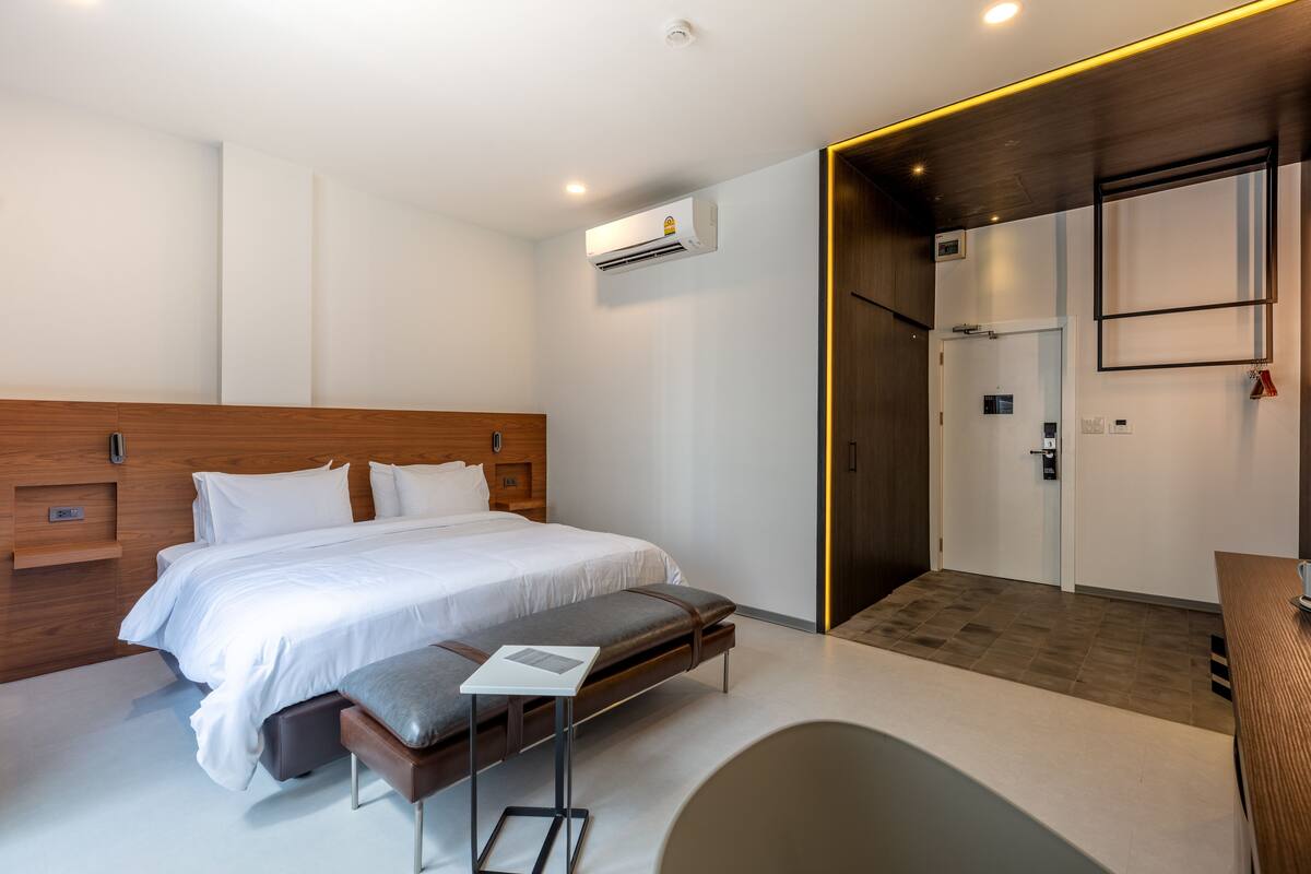 A modern hotel room with a large bed, a bench, a small table, and a walk-in closet.
