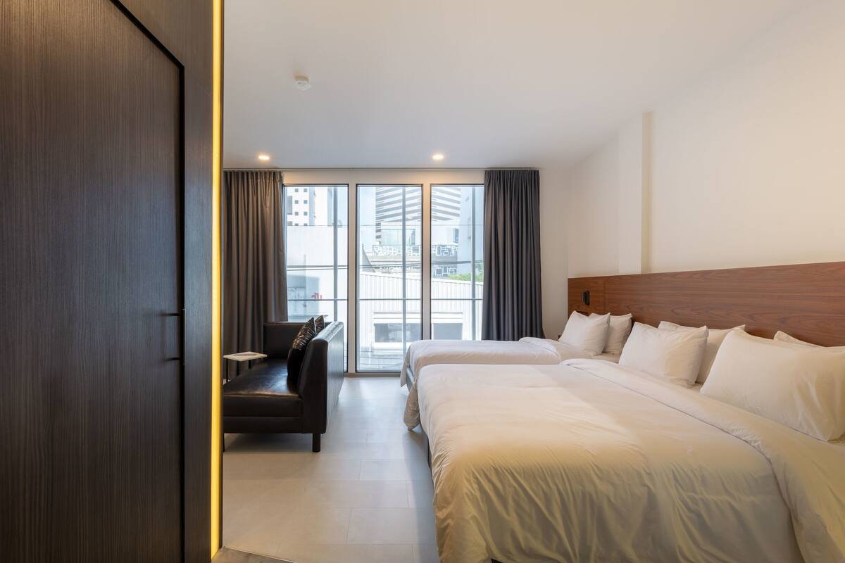 A modern hotel room with a large bed, a sofa, and a view of the city through floor-to-ceiling windows.