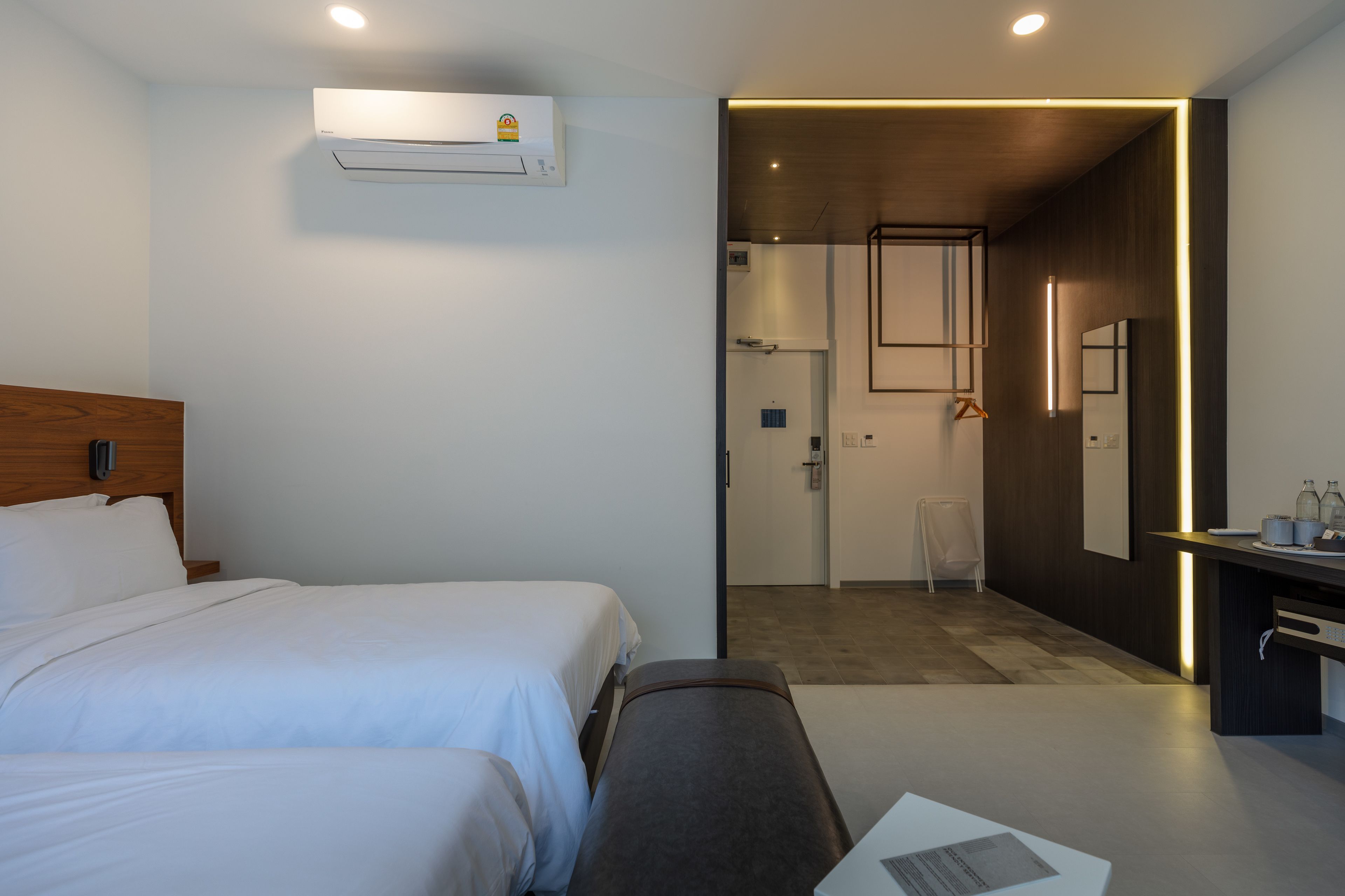 A hotel room with two beds, a bench, a desk with a mirror, and an air conditioning unit.