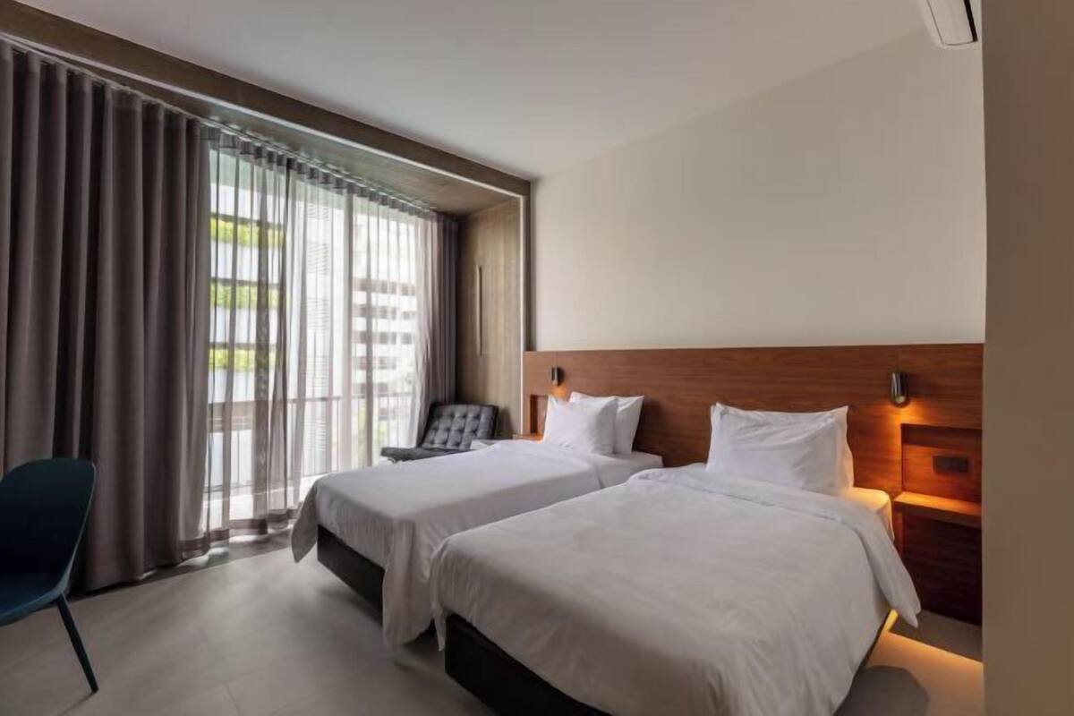 A hotel room with two beds, a chair, a wooden headboard, and a window with curtains.