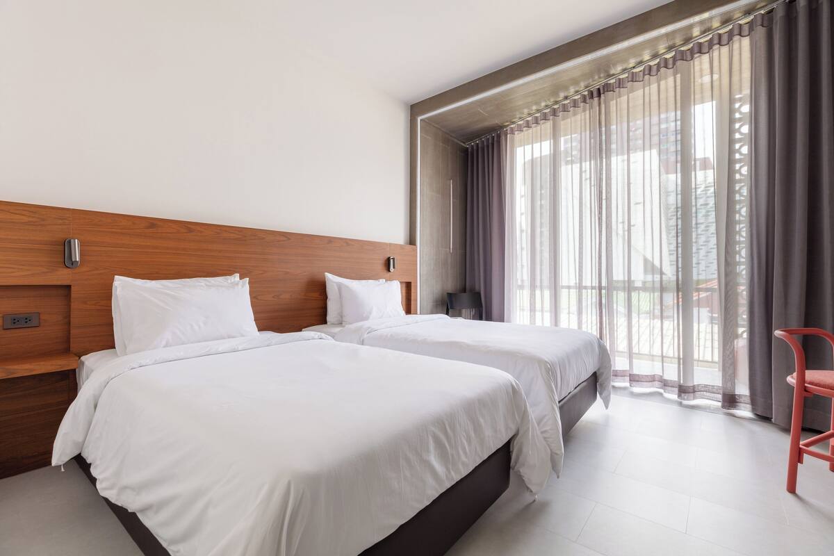 A hotel room with two beds, a wooden headboard, a red chair, and a large window with curtains.