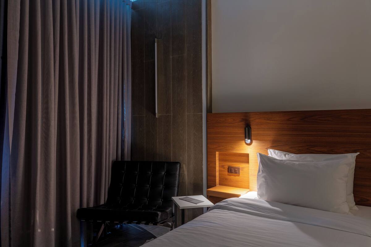 A hotel room with a bed, a black leather chair, a wooden headboard, and a nightstand with a lamp.