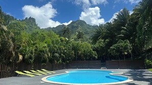 Une piscine dotée d’une terrasse en bois, entourée d’une végétation luxuriante et de palmiers, avec une montagne en toile de fond.