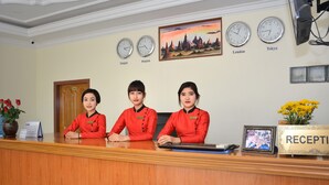 Un comptoir d’accueil avec trois employés en uniforme rouge, des horloges indiquant l’heure à Bangkok, Bangkok et Tokyo, et un tableau représentant un paysage au coucher du soleil.