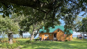 Une cabane en bois avec un toit vert, entourée d’une pelouse bien entretenue et d’arbres, et offrant une vue sur une étendue d’eau.