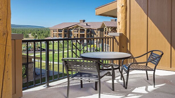 A balcony with a table and two chairs, overlooking a building and a green landscape.