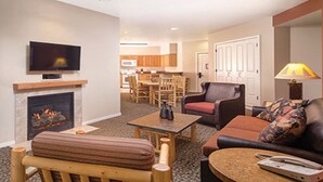 A living room with a fireplace, a television, a sofa, a coffee table, and a dining area in the background.