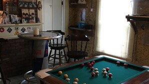 A pool table in a room with a brick wall, a clock, and a window with blinds. - Brookside House-Large, charming Historic Home. Pet Friendly! (Greenwood)