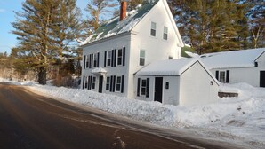 A two-story white house with a green roof, surrounded by snow-covered ground and trees. - Brookside House-Large, charming Historic Home. Pet Friendly! (Greenwood)
