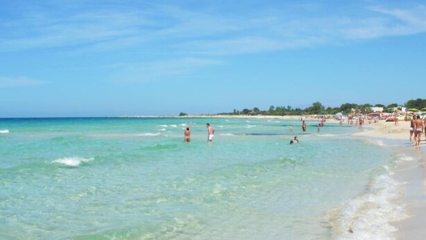 Ein Strand mit klarem türkisblauem Wasser, Menschen beim Schwimmen und Spazierengehen am Ufer, und eine entfernte Küstenlinie mit Gebäuden und Bäumen.