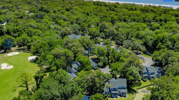 An aerial view of a residential area with houses surrounded by dense greenery, a golf course, and a beach in the background.