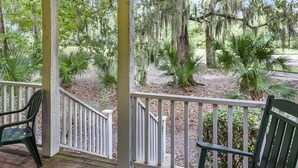 A covered porch with white railings and wooden flooring, overlooking a garden with various plants and trees.