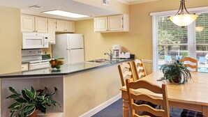 A kitchen with white appliances, a dining area with wooden chairs, and a window with blinds.