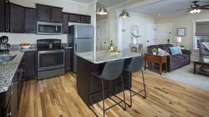 A modern kitchen with dark cabinets, stainless steel appliances, and a granite countertop. There is a bar area with two stools and a small table. The kitchen connects to a living area with a sofa and a coffee table.