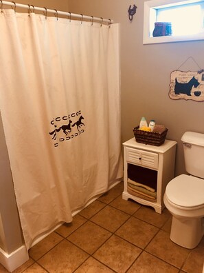A bathroom with a white shower curtain featuring a horse silhouette and Japanese text, a small bedside table with toiletries, and a framed wall decoration.