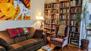 Una sala de estar acogedora con un sofá marrón, una mesita auxiliar, una lámpara, un sillón y una estantería llena de libros.