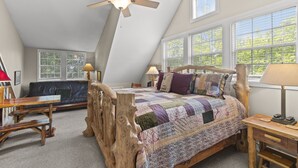 A bedroom with a sloped ceiling, a wooden bed frame, a quilted bedspread, a desk with a lamp, and a window with blinds.